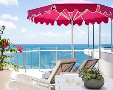 A penthouse apartment balcony with ocean views, a pink beach umbrella and white outdoor lounge and dining furniture.
