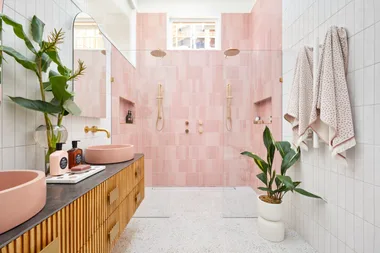 Luxurious bathroom with pink tiles, dual shower heads, pink basins, a wooden vanity, and lush green plants.