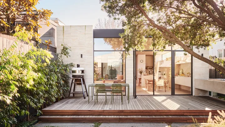 An outdoor kitchen and dining area with a freestanding pizza oven to the left. The back of the house has glass doors and an indoor kitchen is visible through these. Lush greenery is visible, growing beyond the deck where the outdoor kitchen and dining area is.