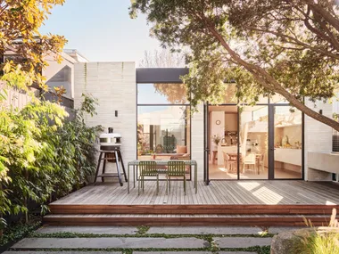 An outdoor kitchen and dining area with a freestanding pizza oven to the left. The back of the house has glass doors and an indoor kitchen is visible through these. Lush greenery is visible, growing beyond the deck where the outdoor kitchen and dining area is.