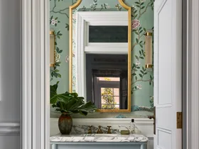 A powder room with wallpaper, wainscoting, a basin built in to a vanity with joinery and a brass-framed mirror.