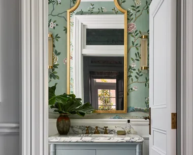 A powder room with wallpaper, wainscoting, a basin built in to a vanity with joinery and a brass-framed mirror.