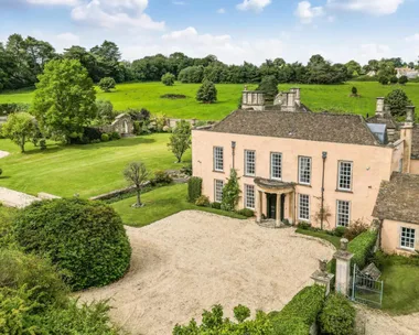 A grand stone manor house, built in 16th Century style with a Queen Anne front pillared portico. Grounds surround the home with trees, hedges, bushes and lush grass. This house was the filming location of the Bennet family house in the BBC adaptation of Jane Austen's Pride and Prejudice.
