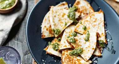 Quesadillas on a dark plate topped with green sauce, next to a drink with lime and ice.