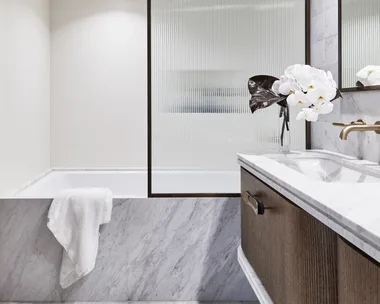 Ensuite bathroom with tub, wooden vanity, white towel, and vase of white flowers. The bathtub is built in and the bath fascia is marble, as is the flooring and the vanity benchtop. A shower screen is framed in black and the timber vanity has brass handles.