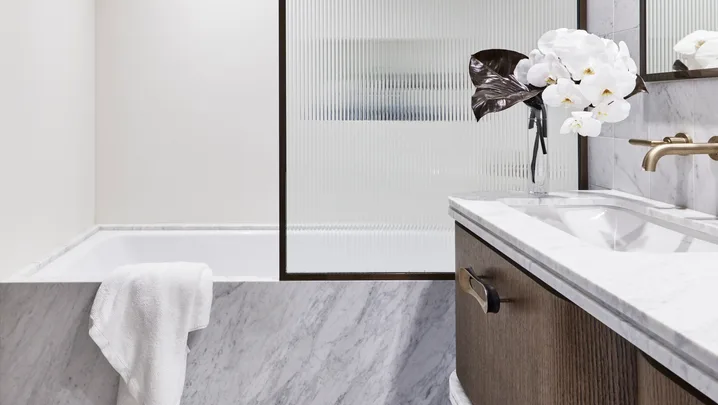 Ensuite bathroom with tub, wooden vanity, white towel, and vase of white flowers. The bathtub is built in and the bath fascia is marble, as is the flooring and the vanity benchtop. A shower screen is framed in black and the timber vanity has brass handles.