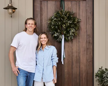Kirsten and Rhys Stanley in front of their front door. The home is a modern farmhouse style with stone cladding, VJ panelling and a wooden door. A wreath for Christmas decorates the door.