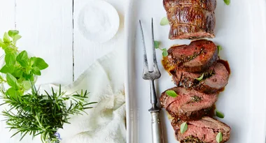 Sliced roasted beef tenderloin with herbs on a white platter, accompanied by fresh rosemary and oregano sprigs.