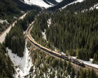 Aerial view of a train traveling through a snowy mountain landscape with pine forests and a towering peak in the background.