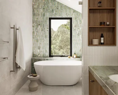 An ensuite bathroom. A vanity is on the right side and in the background is a feature wall with sage green tiles in an arch shape. A window looks on to vistas of bushland behind a curved, freestanding bath.