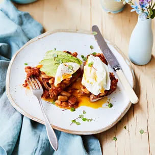 Avocado toast with tomato beans, poached eggs, and herbs on a white plate with a fork and knife.