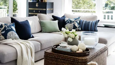 Cozy living room with a beige sectional sofa, patterned cushions, a throw blanket, wicker coffee table, and decorative items.