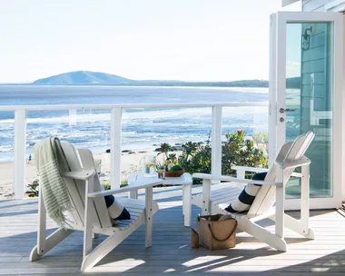 Two white chairs on a beachside deck with a view of the ocean and mountains; glass doors open to a sunny scene.
