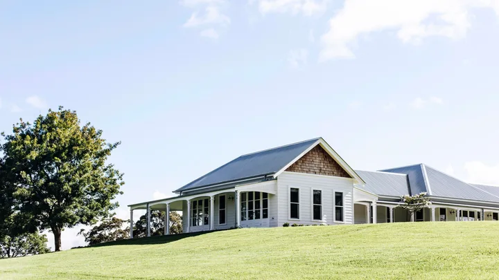 Single-story white house with a gray metal roof, surrounded by green lawn and a tree, under a blue sky.