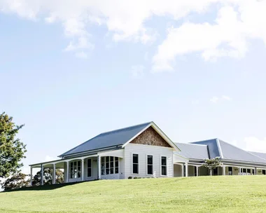 Single-story white house with a gray metal roof, surrounded by green lawn and a tree, under a blue sky.