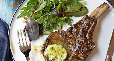 Grilled steak with herb butter and fresh greens on a white plate, with a fork and knife on the side.