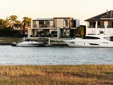 Modern canalside house with large glass windows, palm trees, and two yachts moored by the dock at sunset.