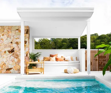Outdoor poolside lounge with white seating, tan pillows, stone wall, potted plants, and a rattan side table.