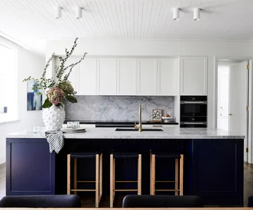 Modern kitchen with a navy island, white cabinetry, marble countertop, large vase with flowers, and three bar stools.