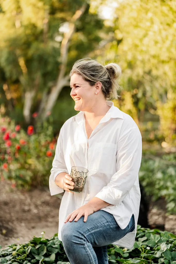 A person in a vegetable garden, smiling and holding a glass, wearing a white shirt and jeans, with trees and flowers in the background.