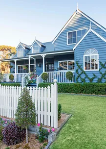 Blue house with white accents, gabled roof, front porch, garden, and white picket fence under a clear sky.