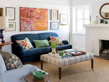 A living room with white walls and ceiling, blue couches on top of a sisal rug and a striped ottoman which doubles as a coffee table. A fireplace has a round mirror above it. The palette feels perfect for winter decorating and could inspire winter decorating ideas.