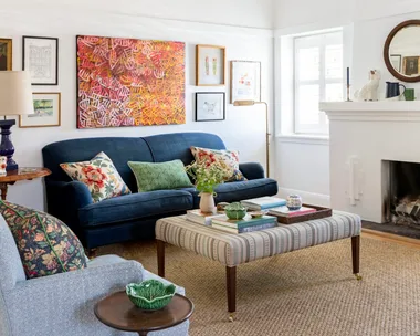 A living room with white walls and ceiling, blue couches on top of a sisal rug and a striped ottoman which doubles as a coffee table. A fireplace has a round mirror above it. The palette feels perfect for winter decorating and could inspire winter decorating ideas.