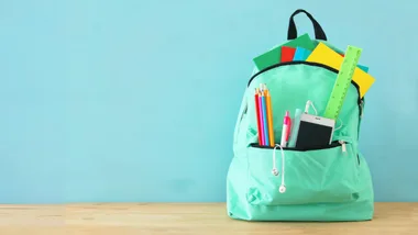 A mint green backpack filled with colorful notebooks, pens, a smartphone, and a ruler against a light blue background.