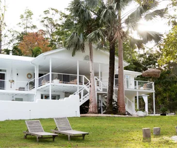 White house with large porch and palm trees, surrounded by greenery; two wooden lounge chairs on the lawn.