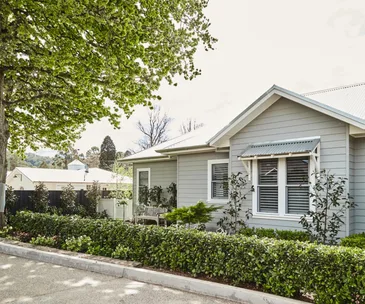 A charming light grey house with white trim, surrounded by lush greenery and a large tree, under a clear sky.